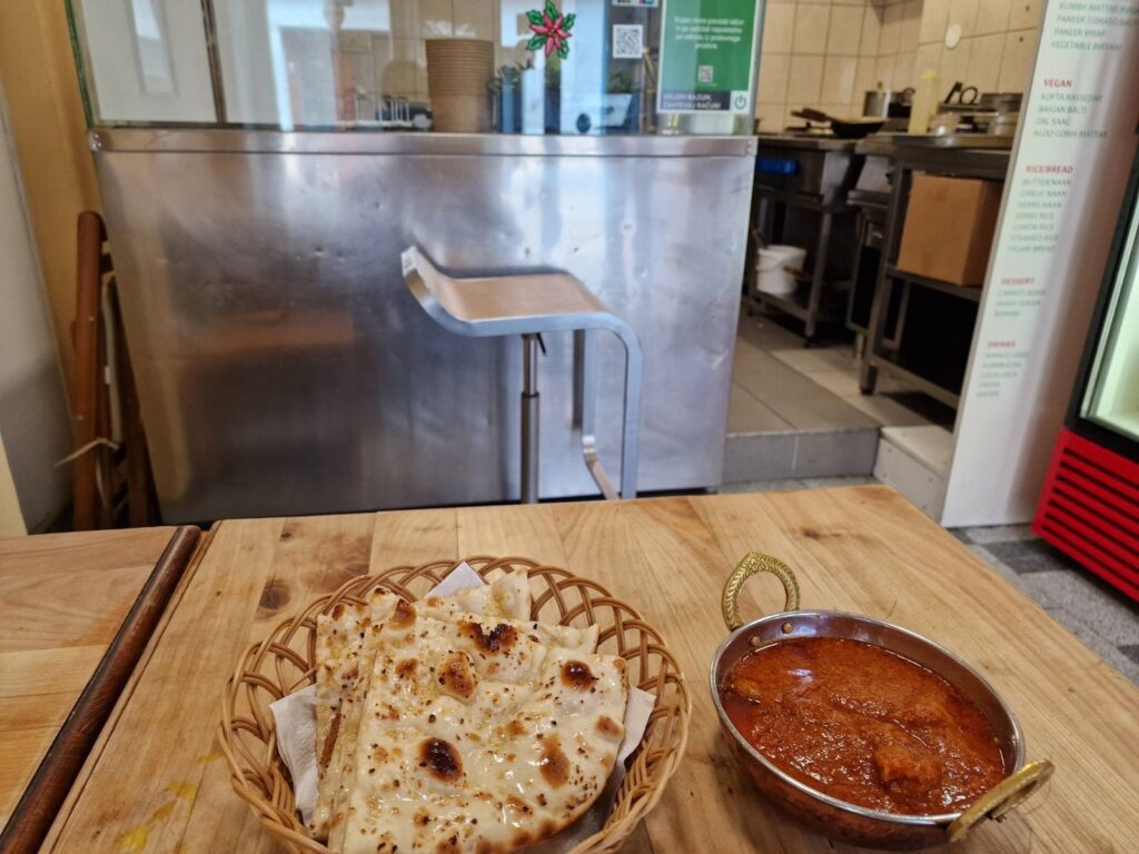 Restaurant kitchen in the background, plate of naan bread to the left and metal round dish with brown-reddish food (sauce) in the foreground
