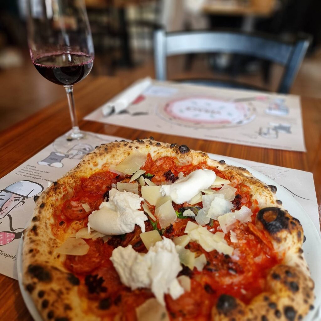 Picture of a glass of red wine in the top left background and pizza on the main frame, individual pizza with burrata and nduja